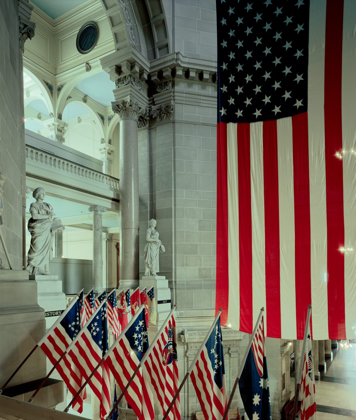 American flags in various historical designs are displayed throughout a classical neoclassical interior with ornate columns, arches, and marble statuary, creating a composition that juxtaposes patriotic symbolism with architectural grandeur.