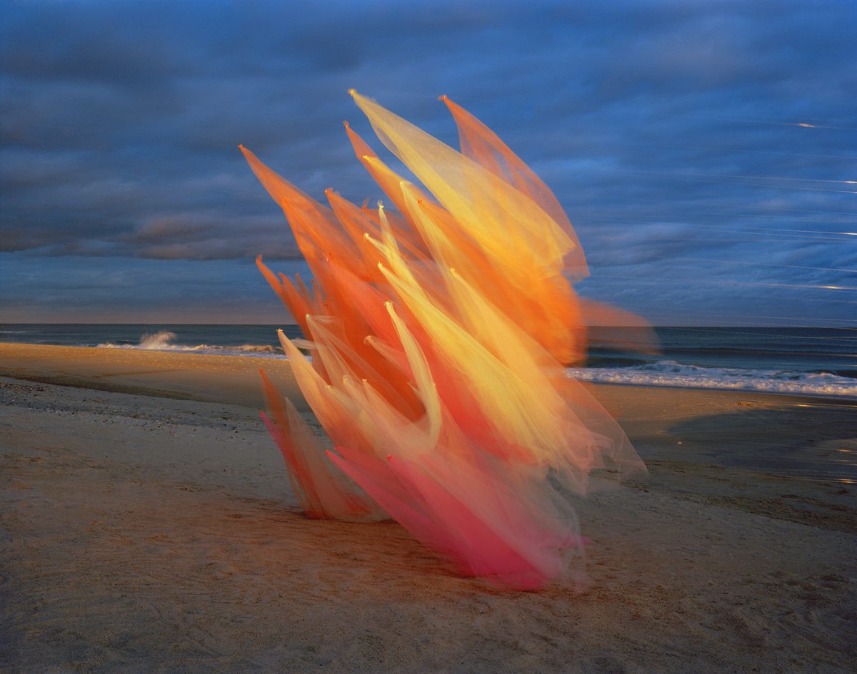 Translucent fabric in graduated shades of coral, orange, and yellow billows on a beach at dusk, creating sharp angular peaks against a blue-grey sky and ocean horizon.