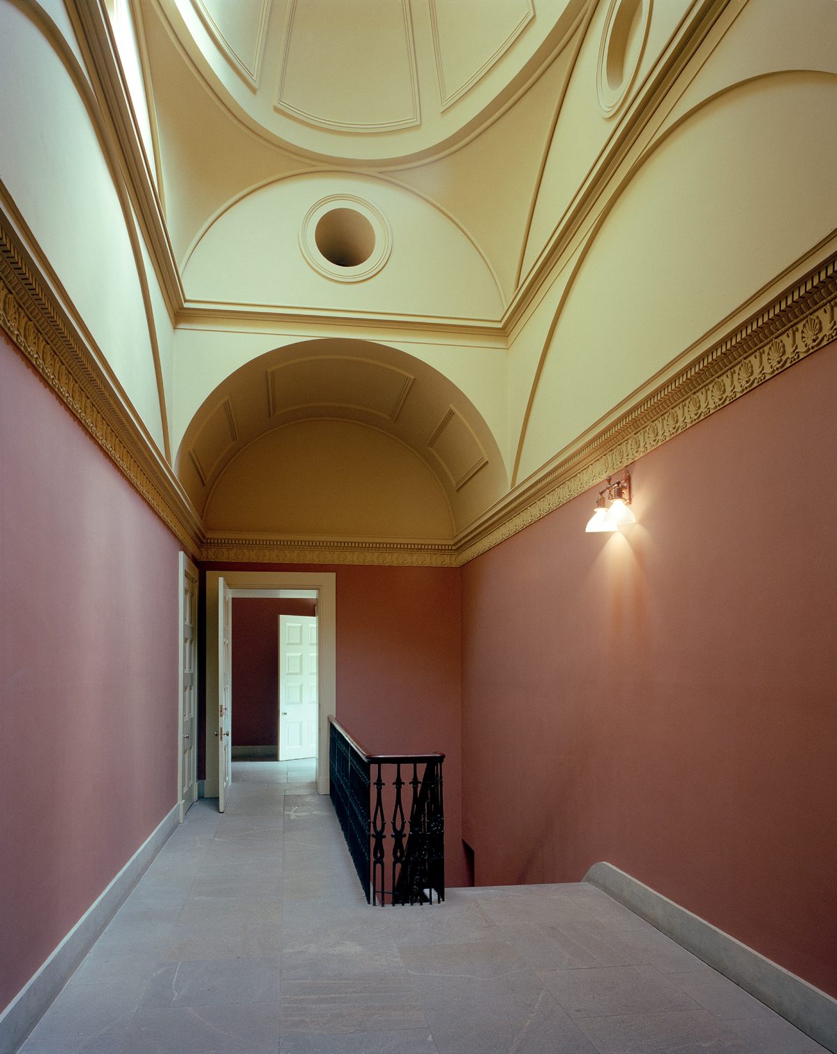 A classical architectural corridor with terracotta-red walls and cream-colored ceiling features ornate molding and a barrel vault. A black wrought-iron railing frames an opening on the right, while a doorway with white trim leads deeper into the space ahead, creating a sense of perspective and geometric symmetry.