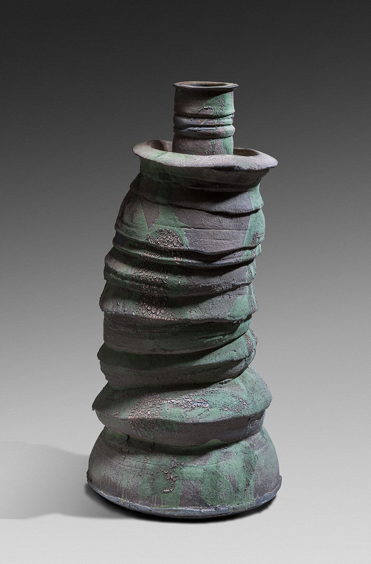 Tall cylindrical ceramic vessel with a conical base, composed of stacked horizontal bands that create a rippled, organic form. The dark stoneware surface is covered with green and gray patina, suggesting oxidation or intentional surface treatment.