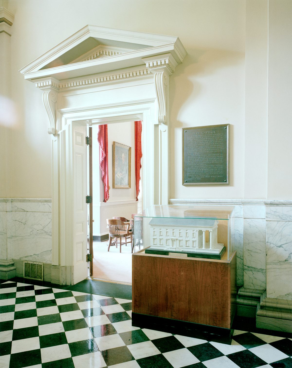 Interior view of a neoclassical Capitol building entrance featuring a grand white doorway with decorative pediment and fluted columns. A wooden display case with a glass top sits in the foreground on a green and white checkered marble floor, with a commemorative plaque mounted on the wall above. The space beyond shows red drapery and period furnishings visible through the open doorway.