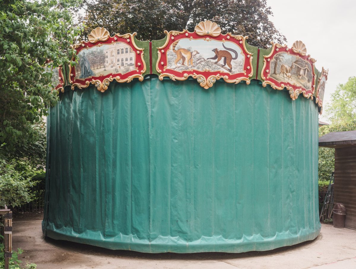 A large cylindrical structure with oxidized copper-green patina is topped with an ornate red and gold decorative carousel-style crown featuring shell motifs and painted panels depicting monkeys and architectural scenes. The structure stands in a wooded outdoor setting.