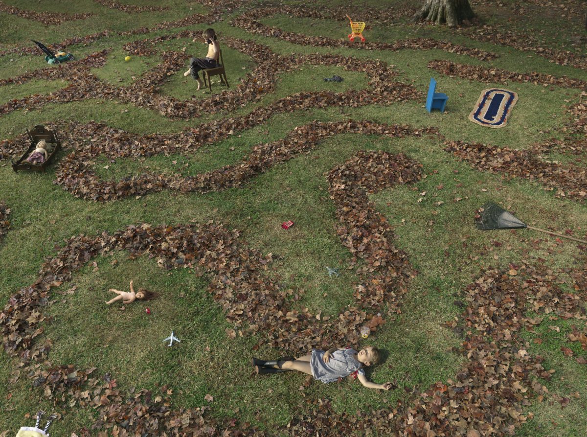 Aerial view of a grassy yard scattered with brown fallen leaves arranged in organic patterns, with two children lying on the grass among the leaves and various toys and objects dispersed throughout the scene, including a chair, rake, and colored play items.