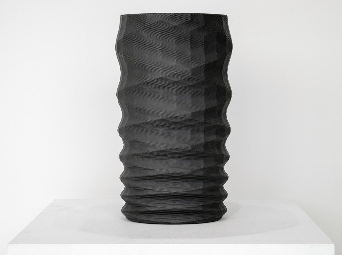 A tall cylindrical black porcelain vessel with undulating, wave-like horizontal ridges along its entire surface, displayed on a white rectangular platform against a neutral background.