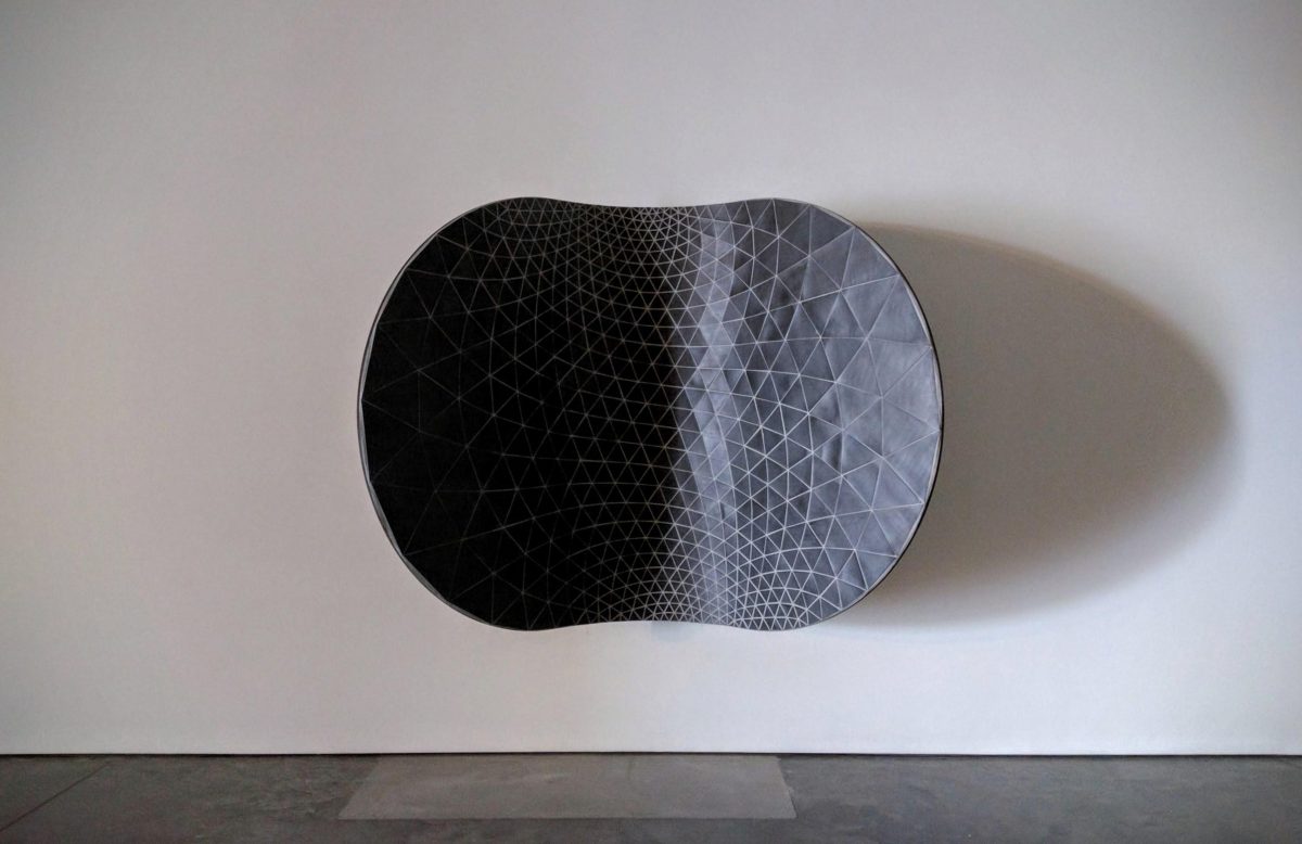 A curved, pod-like sculptural form mounted on a white gallery wall, featuring a densely patterned surface of small triangular tiles that gradient from deep black on the left to light gray on the right, casting a distinct shadow on the wall below.