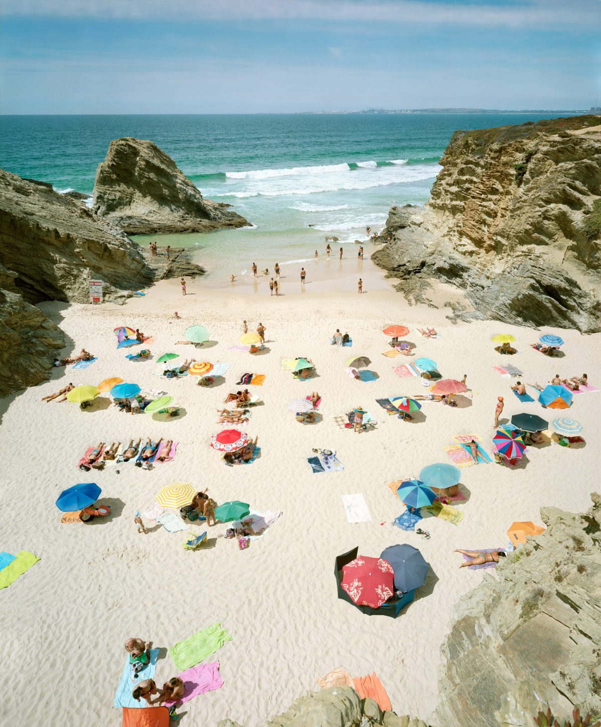 Aerial view of a crowded beach nestled between towering limestone cliffs, with dozens of colorful umbrellas scattered across pale sand and beachgoers swimming in turquoise waters dotted with white waves.
