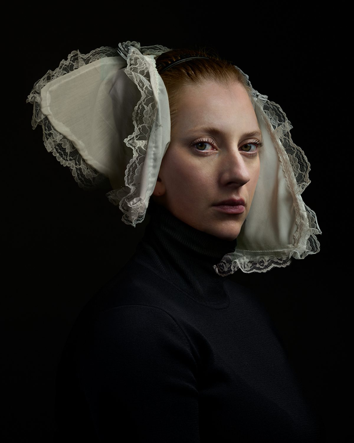 Portrait of a woman in profile against a black background, wearing a black turtleneck and an elaborate white linen bonnet with delicate lace trim and ties. The composition emphasizes the geometric structure of the historical headpiece and the subtle modeling of light across her face and neck.