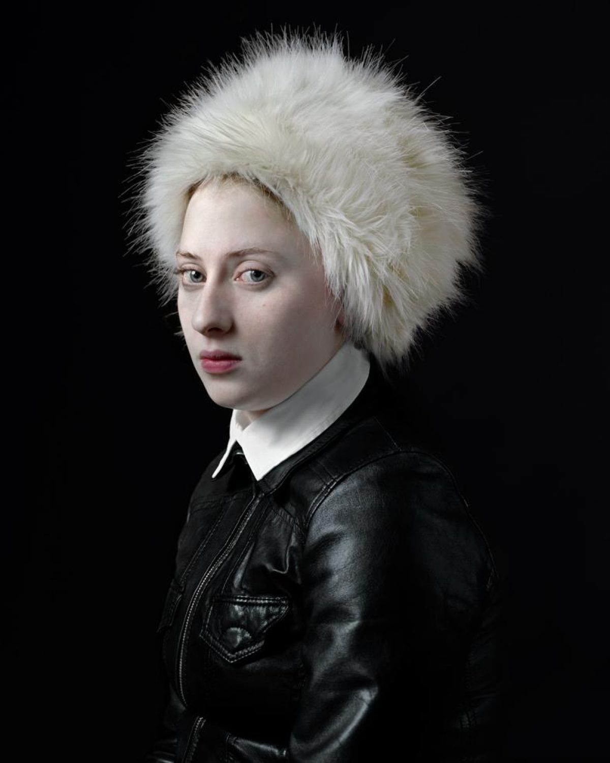 Portrait of a person with pale complexion wearing a voluminous white fur hat and black leather jacket with white collar against a dark background, shown in three-quarter view facing left.