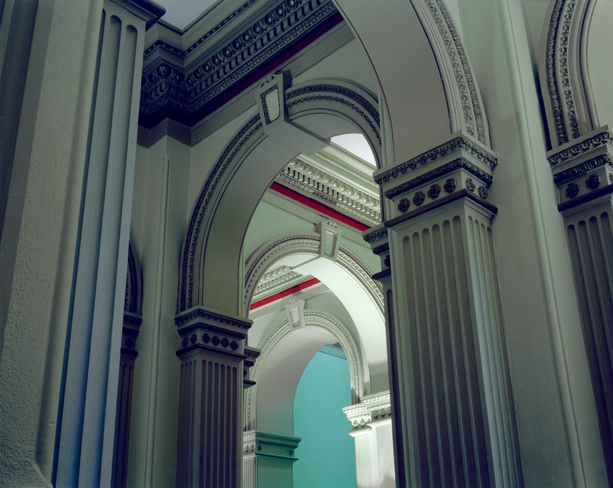 A symmetrical architectural corridor receding into depth, featuring classical arched doorways with ornate moldings and fluted columns in white and dark blue. Glimpses of colored walls—turquoise, red, and green—visible through successive archways, creating a layered perspective down the hallway.