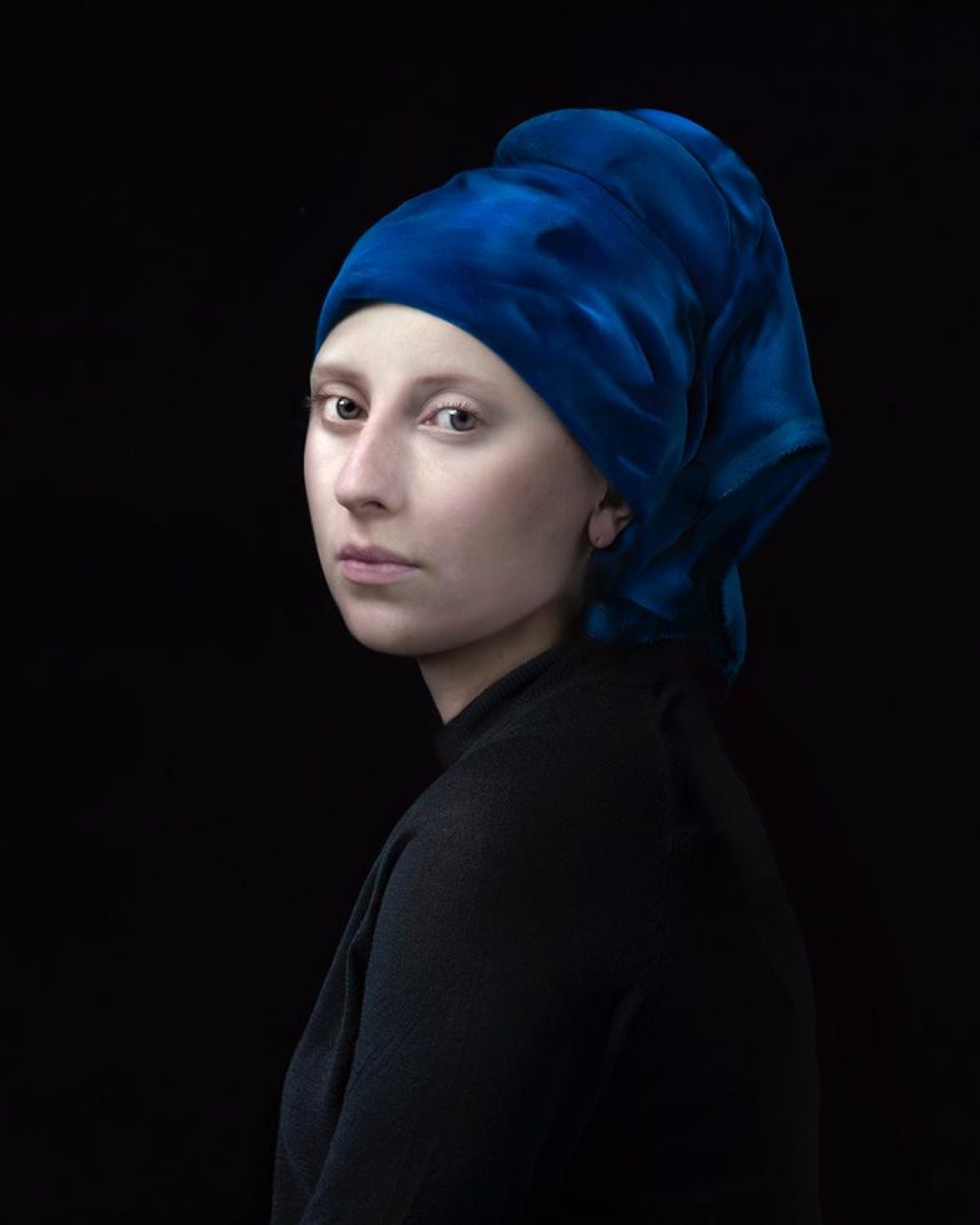 A figure with pale skin wearing a voluminous blue turban and black garment turns to gaze over their shoulder against a dark background, rendered in a contemporary portrait style with striking color contrast and subtle modeling of form.