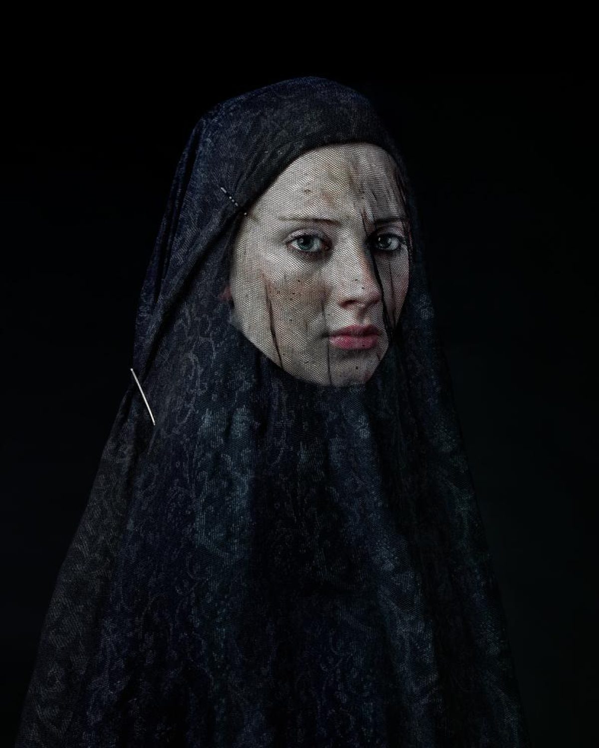 Portrait of a person wearing a dark, textured head covering against a black background. The figure gazes directly toward the camera with a composed expression, their pale face contrasting sharply with the deep navy fabric that drapes over their head and shoulders.