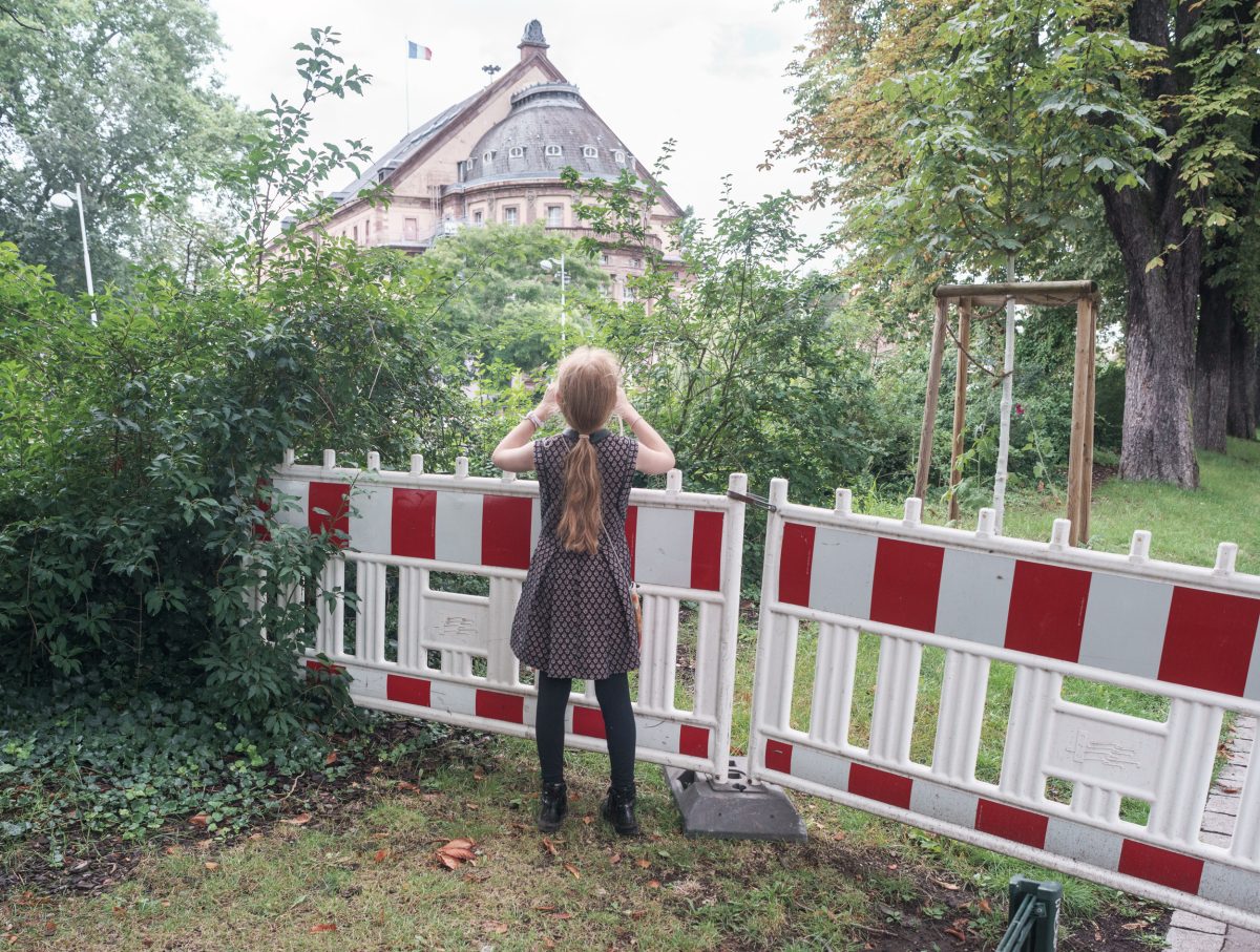A young girl with long hair stands facing away from the camera at a red and white striped barrier in a park setting, her hands raised to her head. Behind her, a distinctive pink neoclassical building with a domed roof and French flag is visible through the trees, creating a striking contrast between the industrial barrier and the architectural landmark.