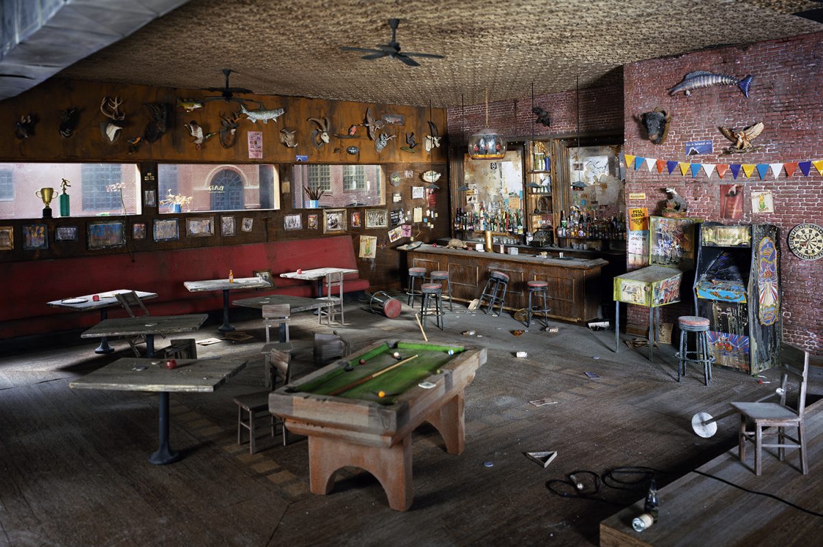 An abandoned interior recreation of a dimly lit bar and recreation room, featuring a pool table in the foreground, a wooden bar counter with shelves of bottles, red vinyl booth seating, taxidermied animal heads mounted on wood-paneled walls, vintage arcade games, and miscellaneous memorabilia scattered throughout. Exposed brick, concrete floors, and ceiling fans create an atmosphere of urban decay and nostalgia.