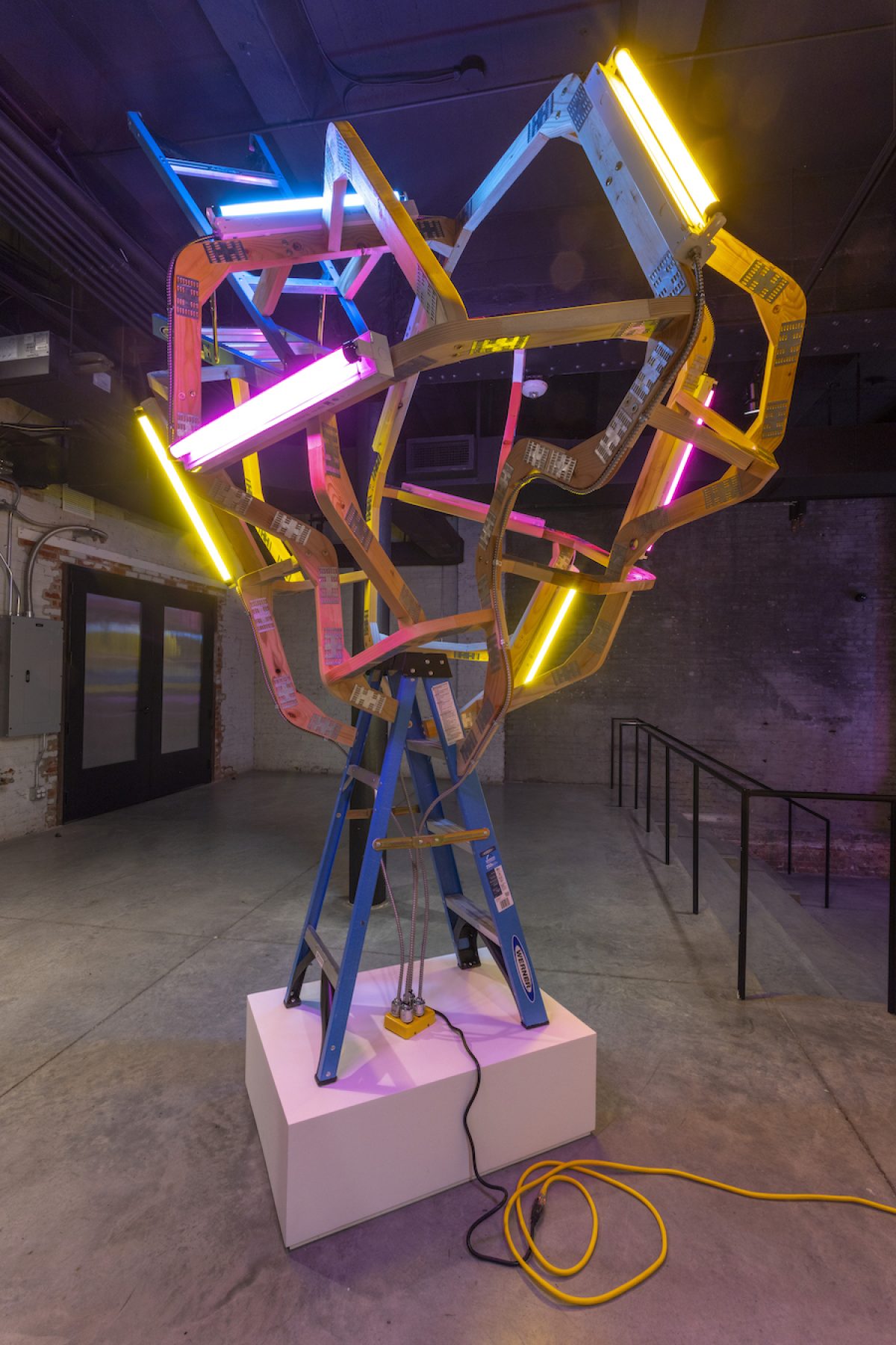 Colorful neon light installation featuring wooden ladders and structural beams arranged vertically on a pink platform. Bright blue, magenta, and yellow fluorescent tubes intersect across the composition, creating an energetic geometric form against the gallery's industrial brick walls.