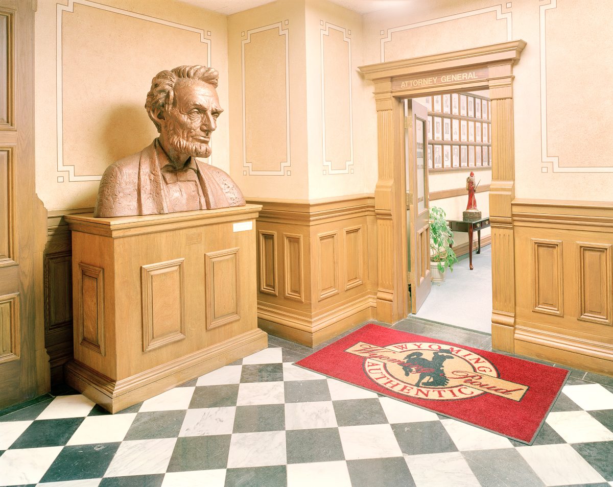 A bronze bust of a bearded historical figure sits on a wooden pedestal in a classical interior hallway with cream-colored walls trimmed in geometric patterns. The space features warm wood paneling, a black-and-white diamond-patterned floor, and an "Attorney General" doorway visible in the background, with a red mat bearing an official seal in the foreground.