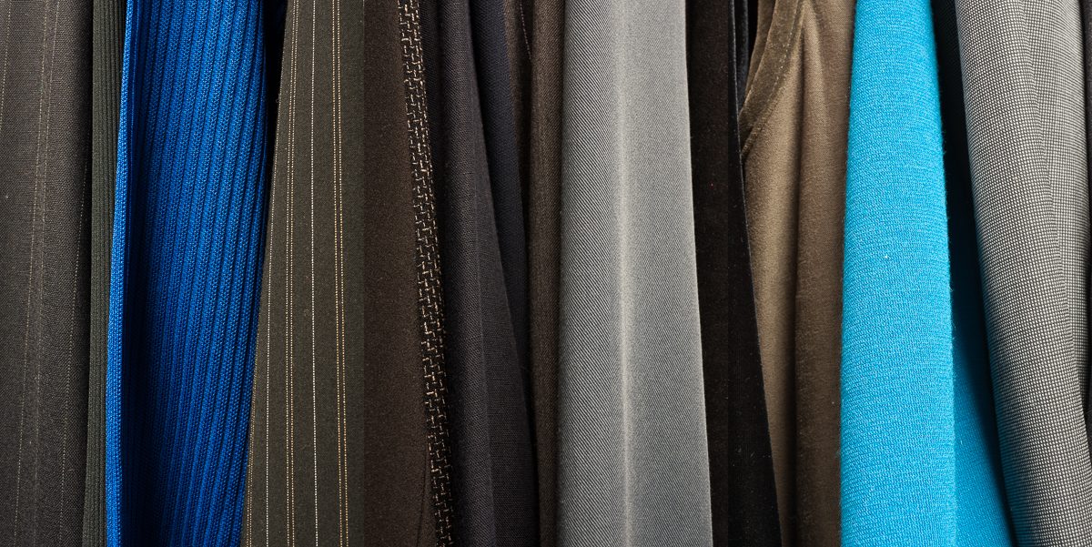 Vertical stripes of fabric in varying textures and colors—charcoal, deep blue, black, gray, tan, and bright cyan—hang closely together creating an abstract composition that emphasizes material contrast and subtle color gradations.