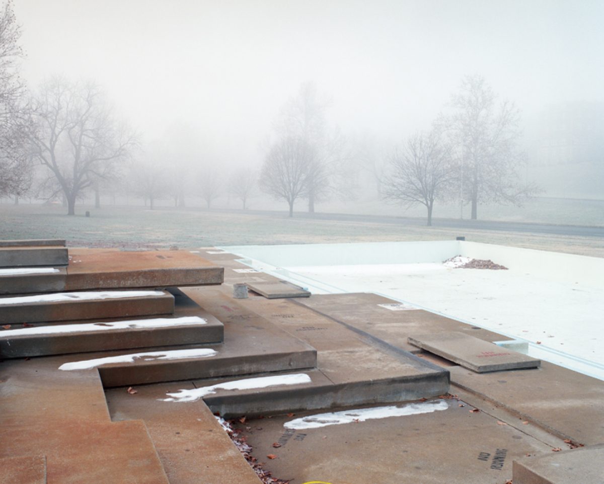 A misty park landscape featuring bare winter trees receding into fog, with a drained concrete wading pool in the foreground. Rusted metal structural elements and wooden beams lie across the pool's floor, creating geometric shadows on the pale surface.