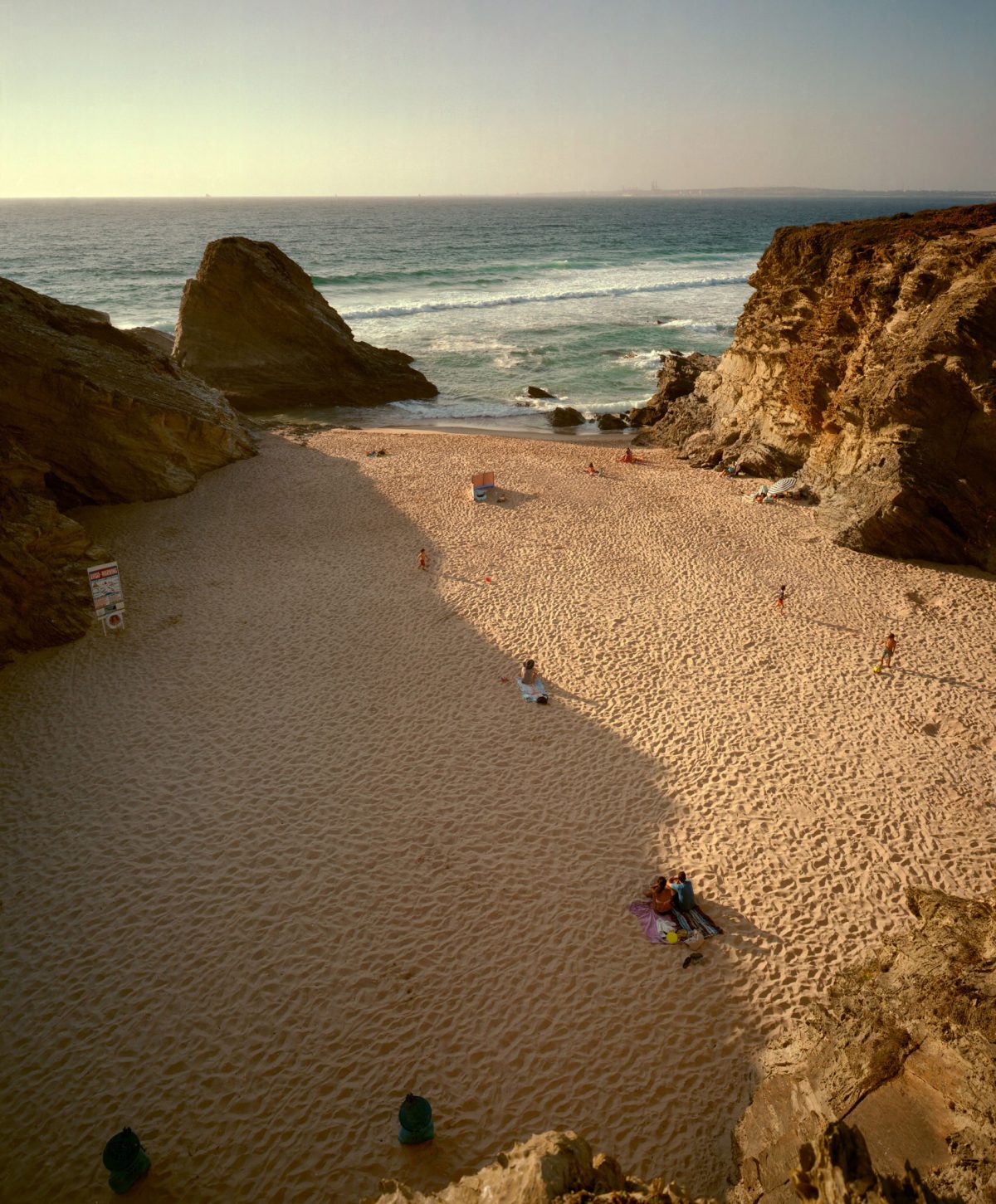 A secluded beach cove framed by towering ochre-colored cliffs, with golden sand and turquoise waters. Several beachgoers are scattered across the expansive shoreline, their small figures emphasizing the dramatic scale of the rocky formations and vast seascape beyond.