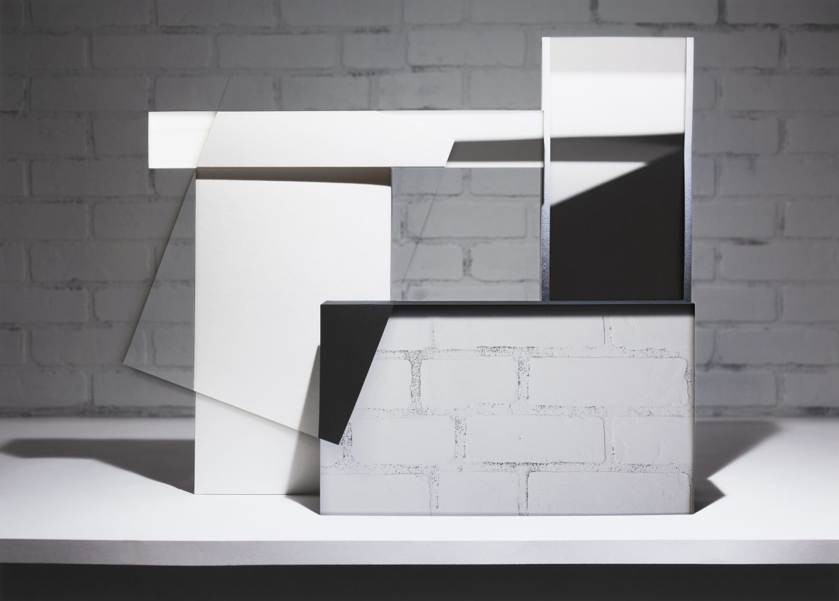 Abstract architectural composition featuring overlapping rectangular planes in white, black, and gray tones arranged in layered, three-dimensional forms against a white brick wall background. Strong diagonal lines and shadow play create depth across the stacked geometric elements.