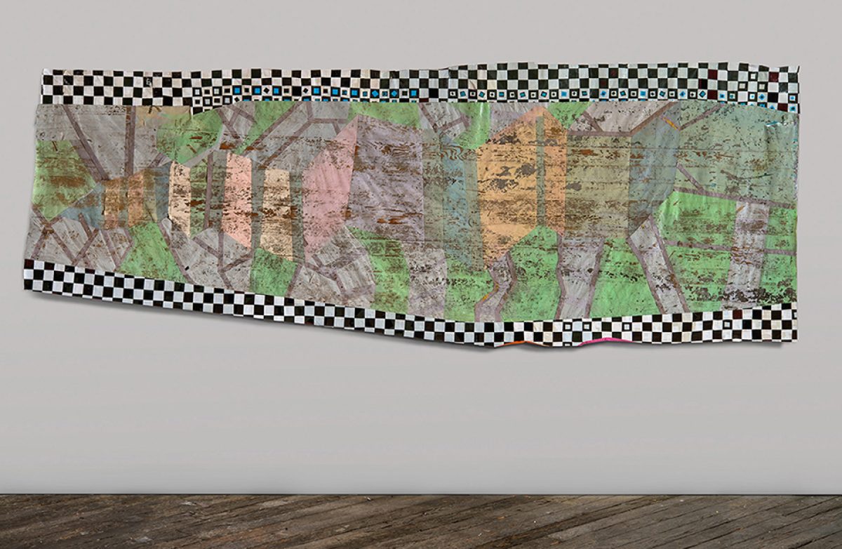A horizontal mixed-media collage featuring layered aerial or cartographic imagery in greens, browns, and muted tones, bordered by a black and white checkered pattern. The composition is mounted on a light gray wall above wooden flooring, with the irregular edges and translucent quality of the materials creating depth and visual complexity.