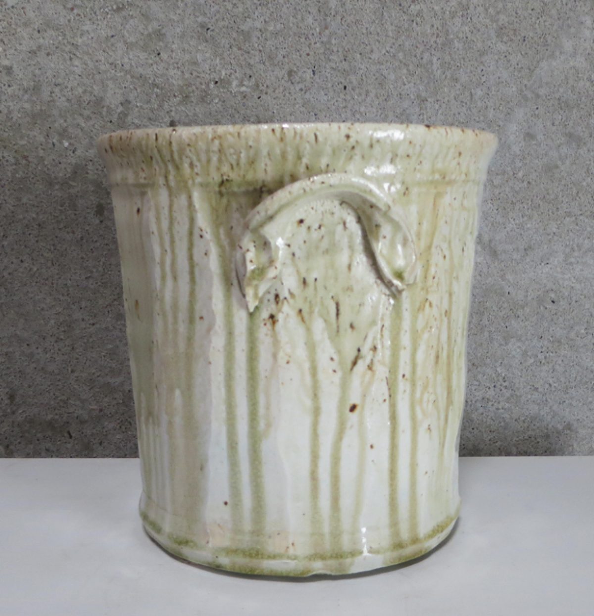 Cylindrical porcelain vessel with a cream-colored glaze featuring vertical striations and organic drip patterns, accented with brown speckles. The piece displays a subtle handle and exhibits the warm, natural variations characteristic of wood-fired ceramics.