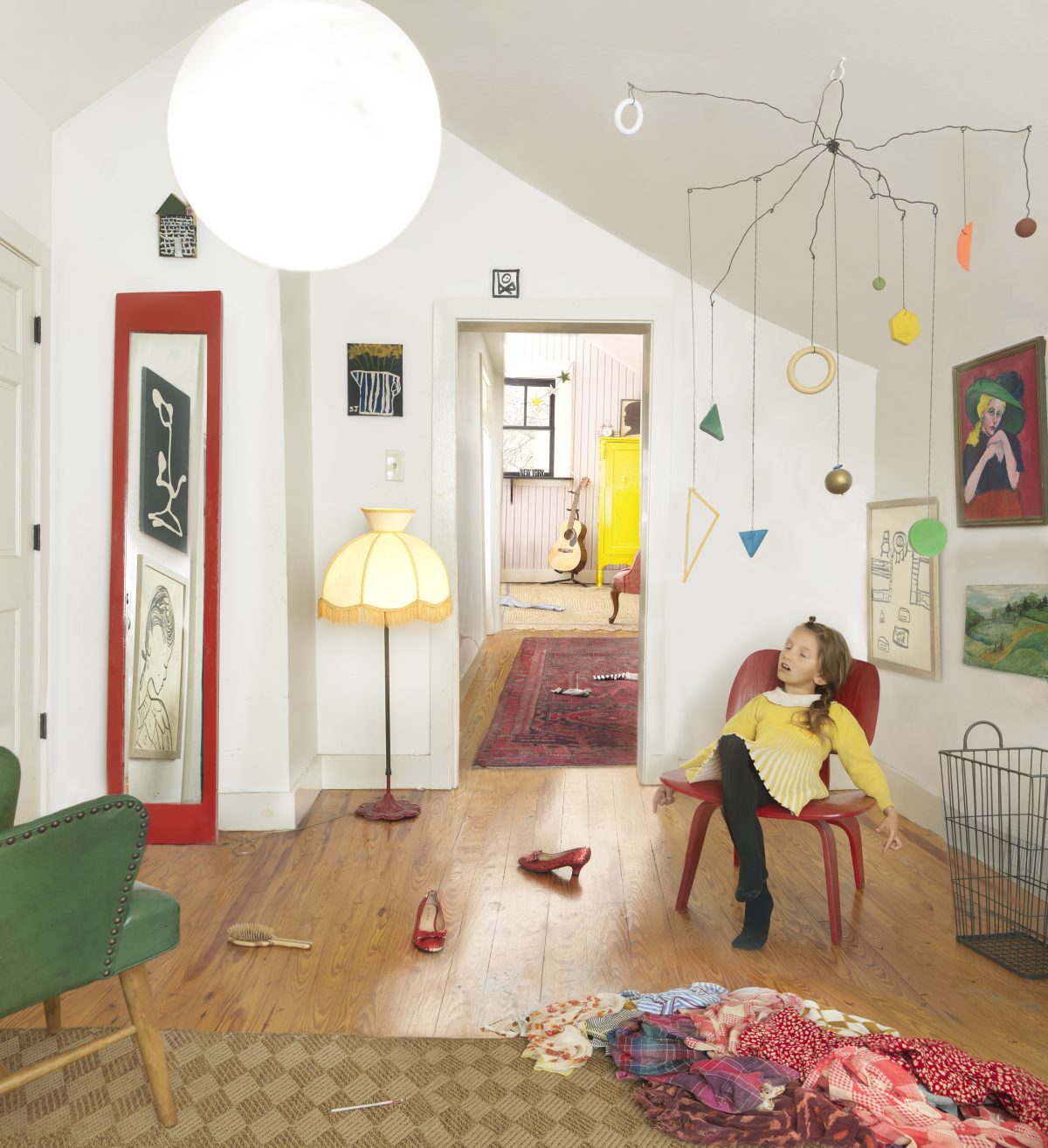 A young girl in a yellow sweater sits in a burgundy chair within a bright, eclectic interior space. The room features white walls, wooden flooring, and is filled with colorful artworks, hanging mobiles with painted geometric shapes and spheres, a yellow-shaded floor lamp, and scattered red shoes on the floor, creating a whimsical, creative domestic environment.