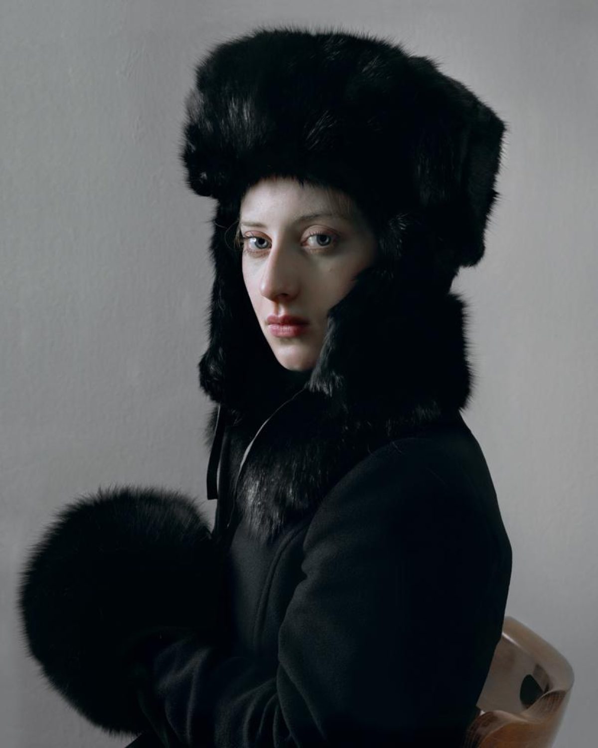 A person with pale complexion gazes over their shoulder while wearing a large black fur hat and collar against a neutral gray background. The subject is dressed in dark clothing with a striking fur muff held at their side, creating a composed portrait with dramatic contrast between the figure's fair skin and the rich black textures of the fur.