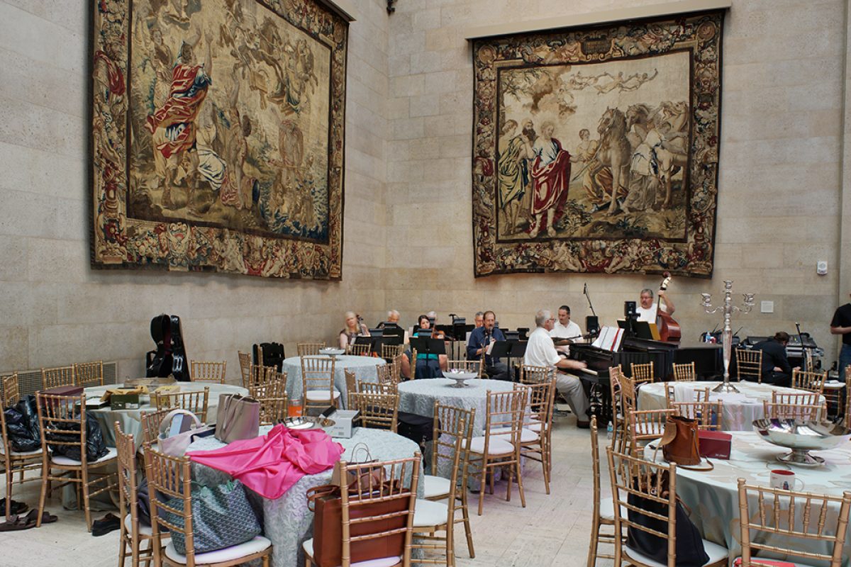 A grand hall with ornate tapestries depicting classical scenes hangs above round dining tables set with white linens, wooden chairs, and colorful fabric accents. A grand piano and musicians are visible in the background as event preparations take place.