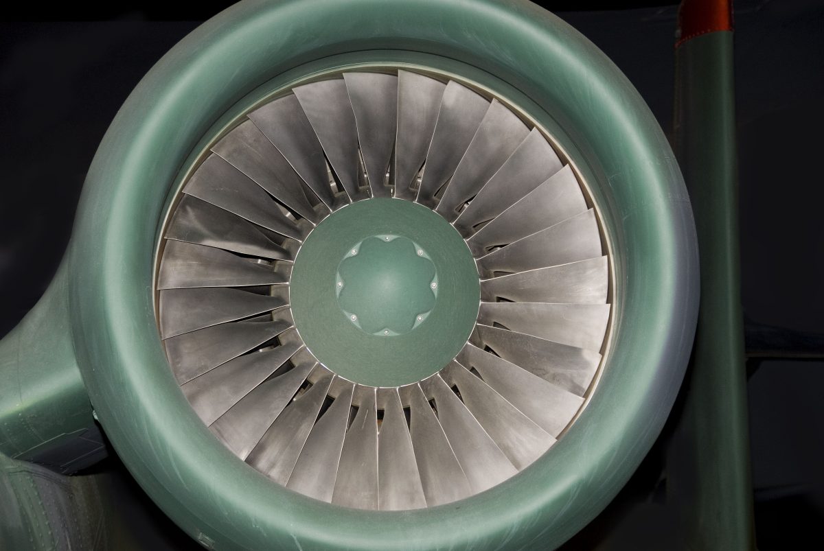 Frontal view of a jet engine intake with concentric metal fan blades radiating from a central green hub, set against a dark background. The polished steel blades create a symmetrical, geometric composition in silvery tones contrasted with the muted sage green of the engine casing.