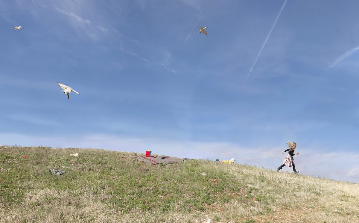 A person in a patterned dress runs across a grassy hillside while multiple kites fly overhead in a bright blue sky, with scattered picnic items visible on the ground below.