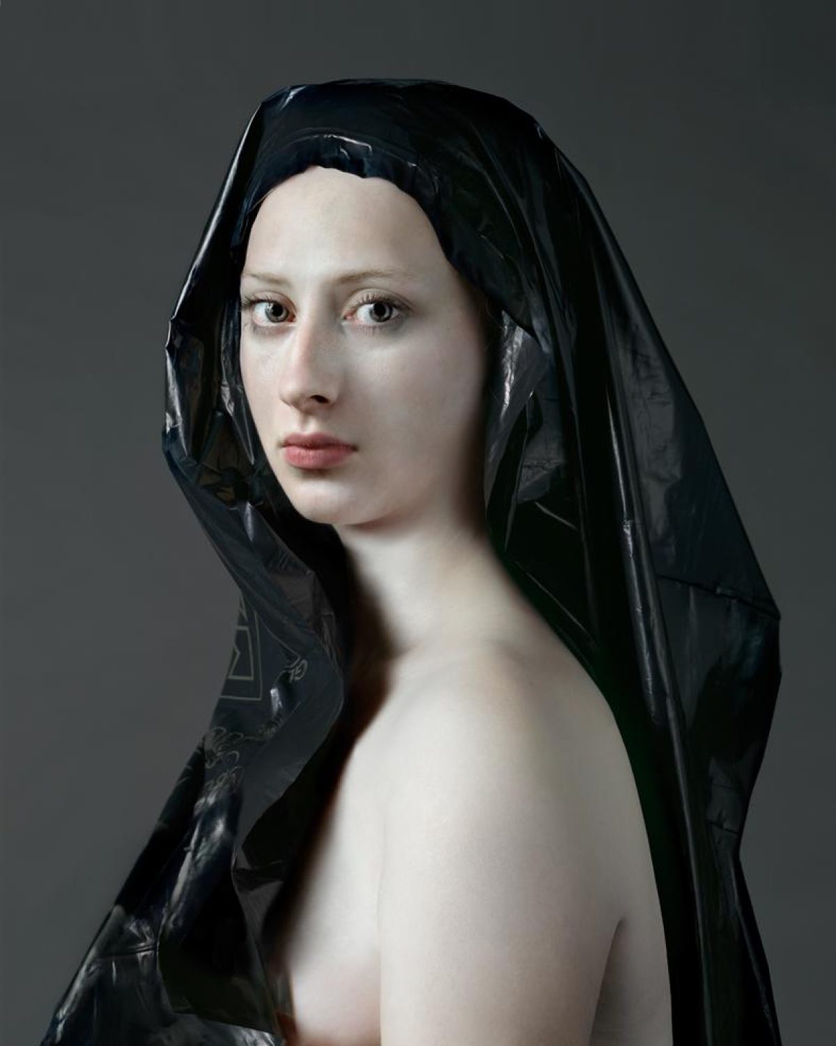 Portrait of a pale-skinned figure with dark hair, wearing a sheer black veil draped over the head and shoulders against a neutral gray background. The composition is a three-quarter view showing refined facial features and bare shoulders, rendered in a classical style with dramatic lighting.