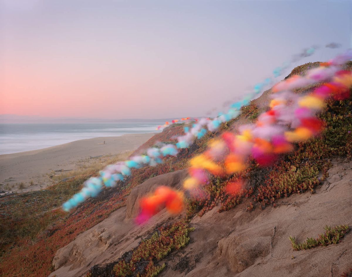 Coastal landscape at dusk featuring a sandy cliff overlooking a beach and ocean, with a diagonal line of vibrant, blurred colored powder or pigment—in shades of cyan, magenta, yellow, and orange—cascading down the hillside against the muted tones of the terrain and twilight sky.