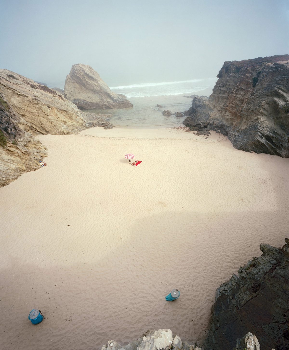 A secluded sandy beach enclosed by dramatic rocky outcrops, viewed from an elevated vantage point. A few colorful beach umbrellas and towels dot the pale sand near the water's edge, while the turquoise sea stretches toward a hazy horizon under an overcast sky.