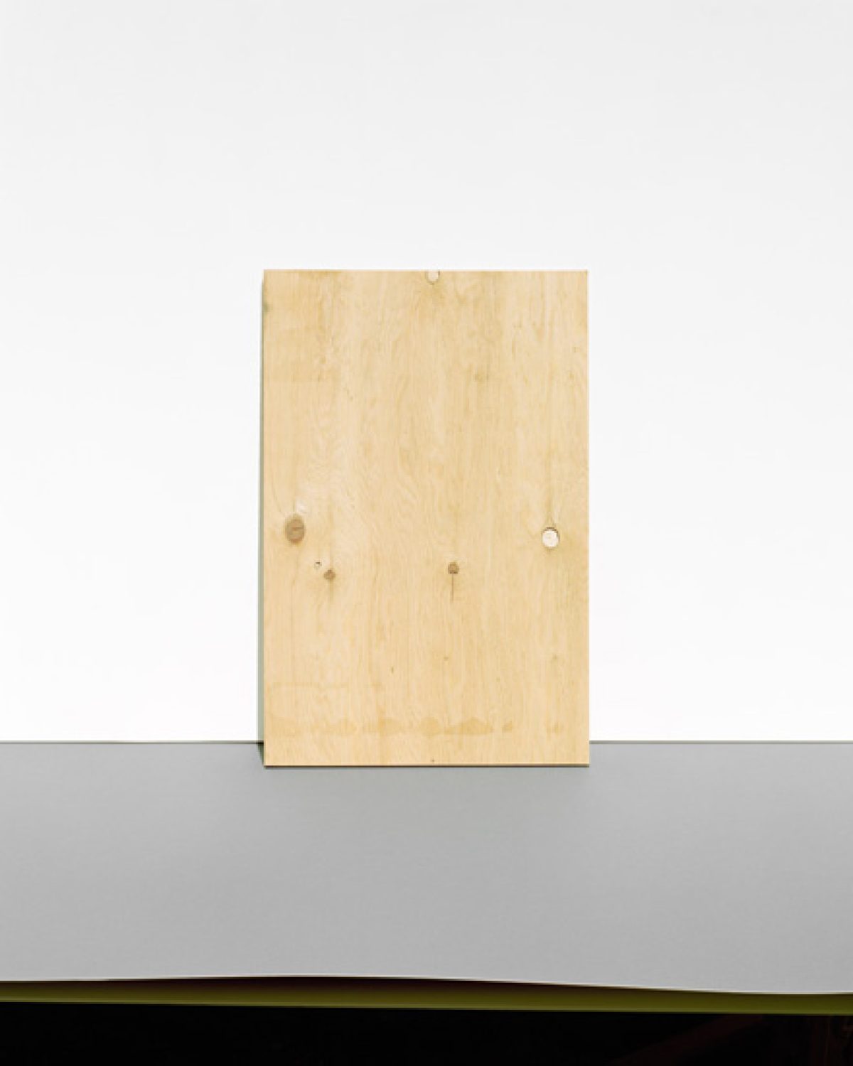 A light wooden rectangular panel leans against a gray wall above a dark horizontal stripe, positioned in a minimalist composition with a plain beige background.