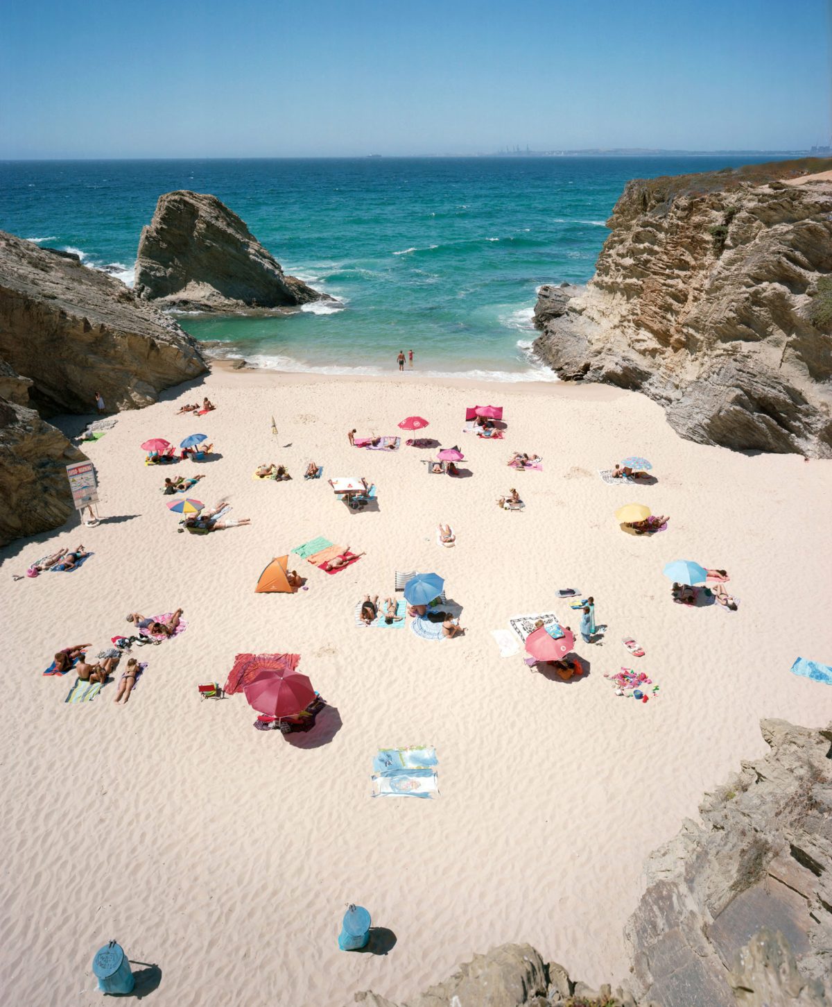 Aerial view of a sandy cove beach flanked by dramatic rocky outcrops, with numerous beachgoers scattered across the sand under colorful umbrellas and beach tents in shades of pink, blue, yellow, and orange. Turquoise water with white-capped waves frames the scene, with a few swimmers visible near the shore.