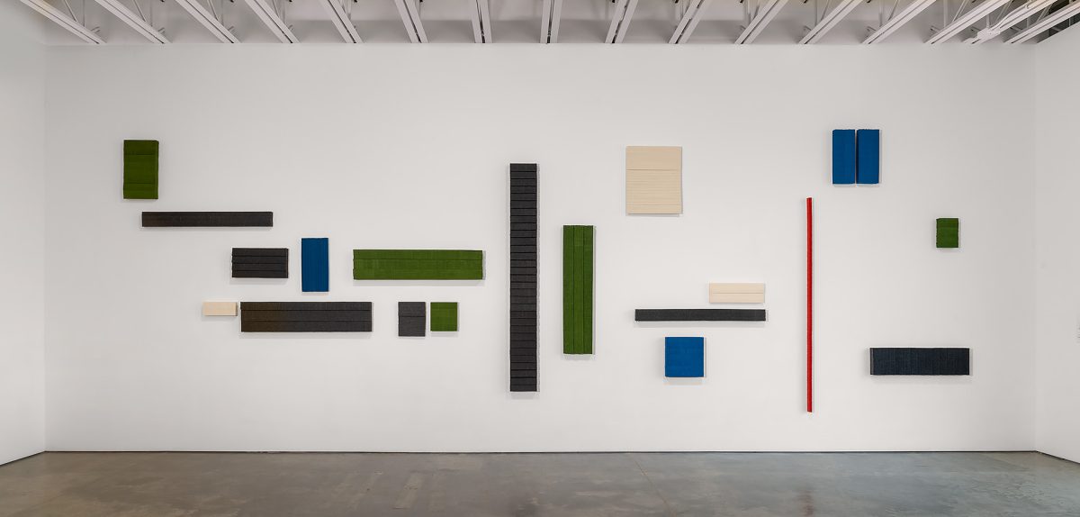 Abstract wall installation composed of rectangular and square panels in varying sizes arranged across a white gallery wall. Panels in dark charcoal, forest green, cream, blue, and a single red vertical element create a rhythmic composition against the minimalist white backdrop.