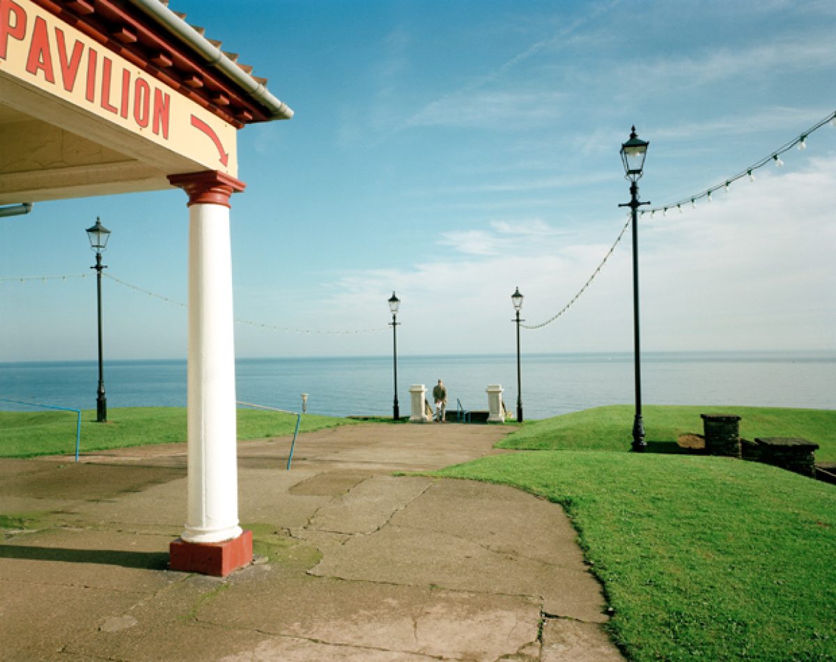 A seaside pavilion with a white column and red trim stands in the foreground, its signage partially visible. Beyond it, manicured green lawns slope toward a calm sea under a pale blue sky, with vintage street lamps and string lights creating a nostalgic atmosphere along the coastal promenade.