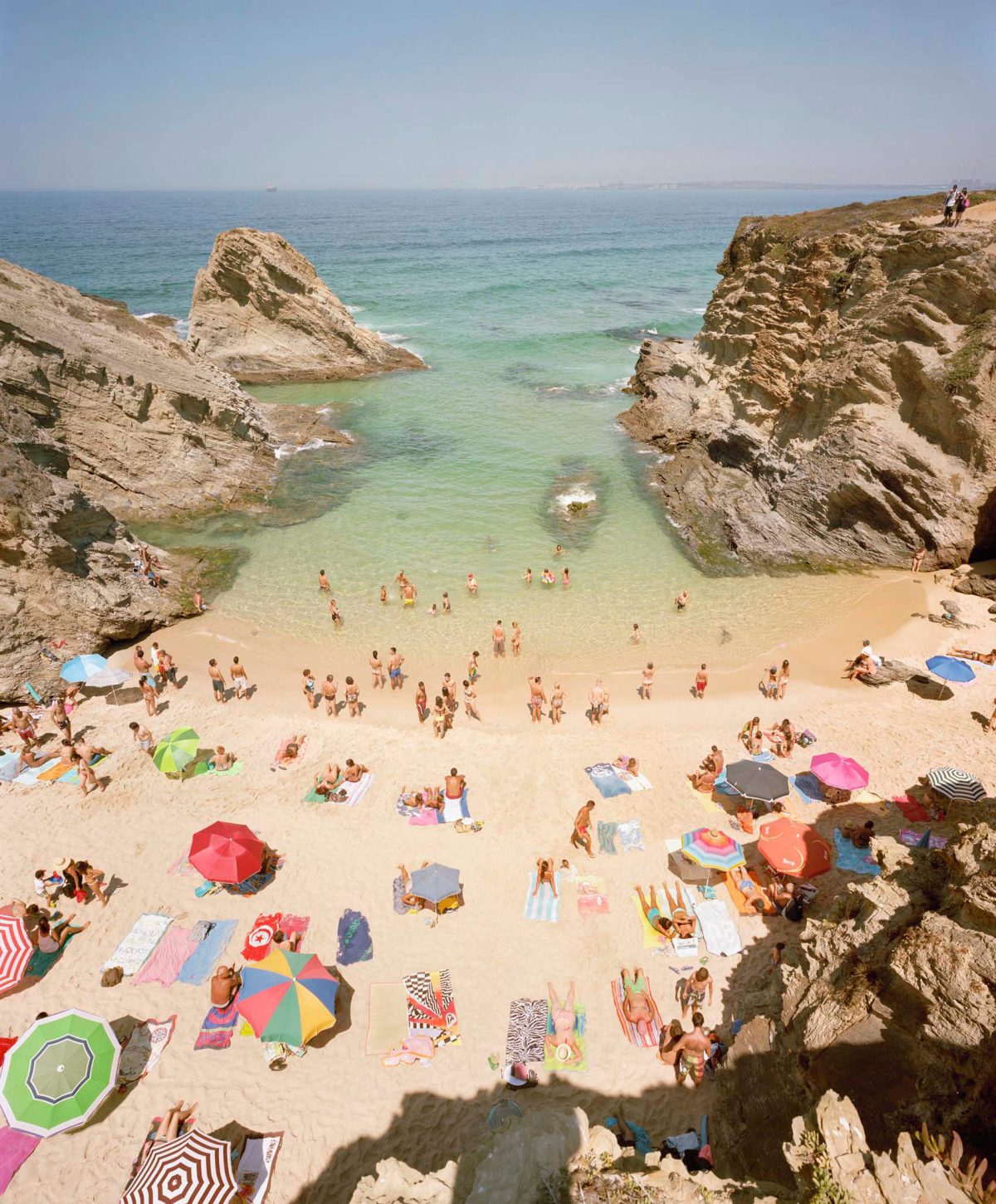 Aerial view of a crowded beach nestled between dramatic limestone cliffs, with colorful umbrellas and beach towels dotting the golden sand, turquoise water, and numerous swimmers and sunbathers scattered across the scene.