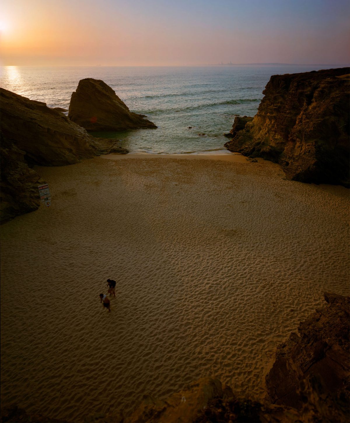 A secluded sandy beach framed by dark rocky outcrops on either side, with a solitary figure walking across the pale sand toward turquoise waters. The sky glows with warm golden and pink tones of sunset or sunrise, creating a luminous reflection on the calm sea horizon.