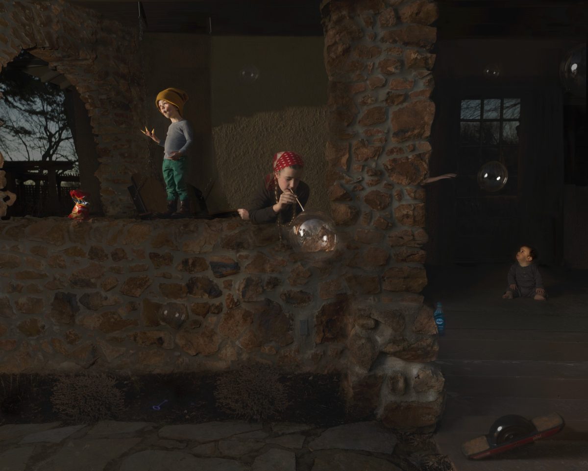 Three children play in a stone-walled interior space illuminated by dramatic natural light. One child in a yellow hat stands to the left, another wearing a red bandana blows soap bubbles in the center, and a third sits near a doorway on the right. The warm, golden light casts strong shadows across the weathered brick and stone surfaces, creating a contemplative atmosphere in this domestic scene.