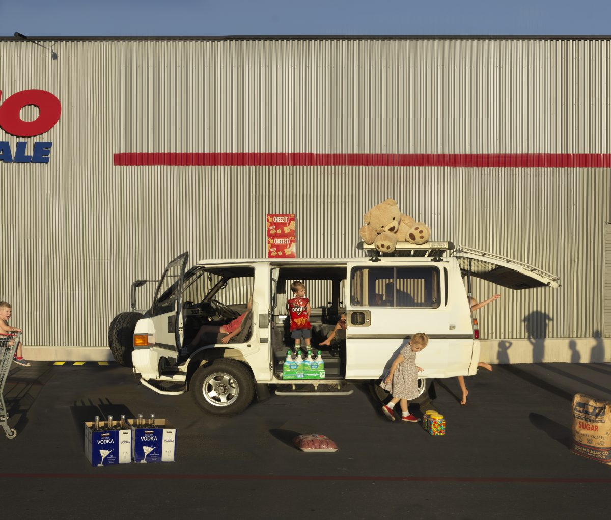 A white minivan loaded with consumer goods and a large teddy bear sits parked in front of an industrial corrugated metal warehouse backdrop bearing a red corporate logo. Children and adults are arranged around the vehicle amid scattered products including vodka bottles, crackers, and a bag of sugar, creating a surreal domestic scene that subverts typical consumer culture imagery through deadpan composition and saturated color.