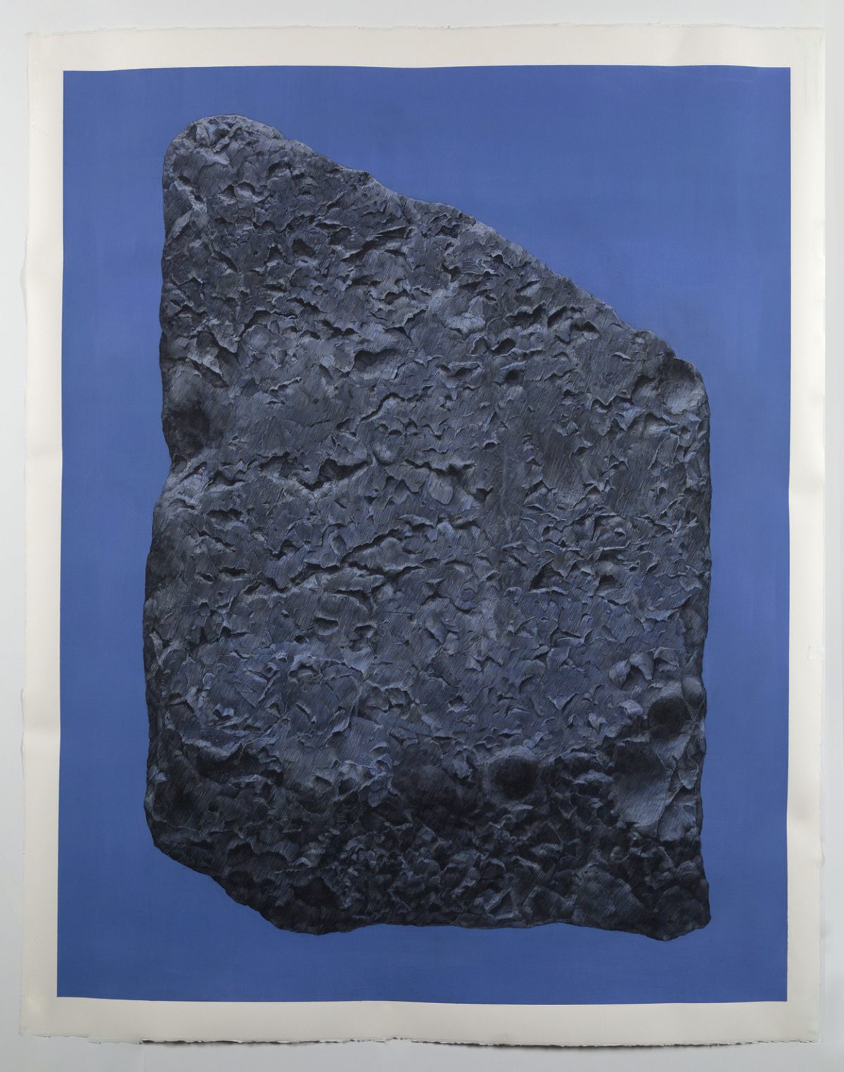 A dark gray textured stone fragment positioned vertically against a deep blue background, its rough surface densely carved with linear marks and patterns that create an intricate, weathered appearance.