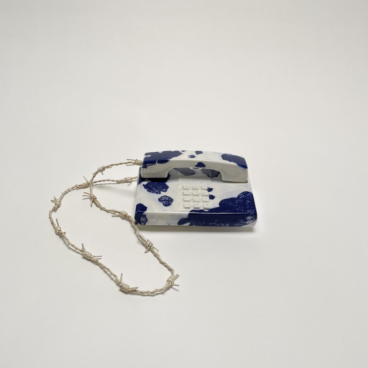 A rectangular porcelain tag with cobalt blue and white cow print pattern, featuring a raised grid of small white dots in the center, suspended from a knotted natural fiber cord.