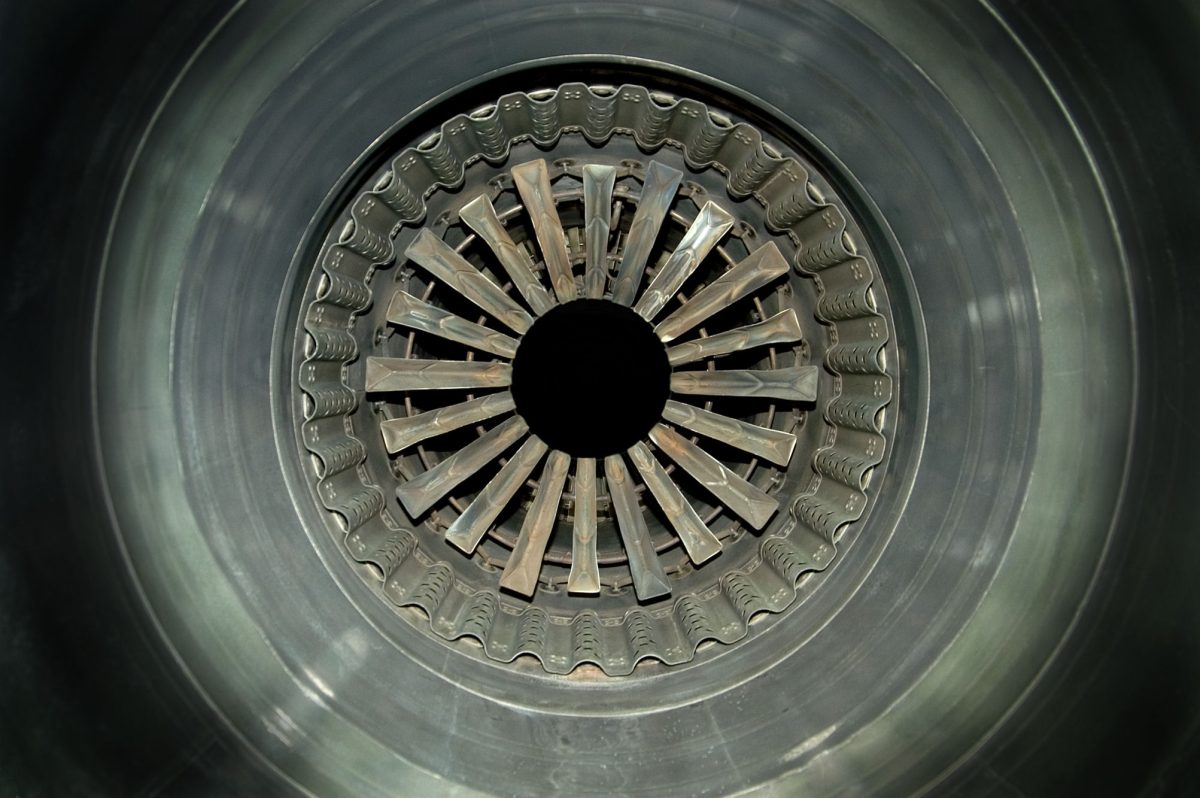 Frontal view of a jet engine intake, showing concentric circles of metallic blades and ducts radiating from a central black aperture. The photograph captures the geometric precision and industrial symmetry of the engine's interior in cool gray and silver tones.