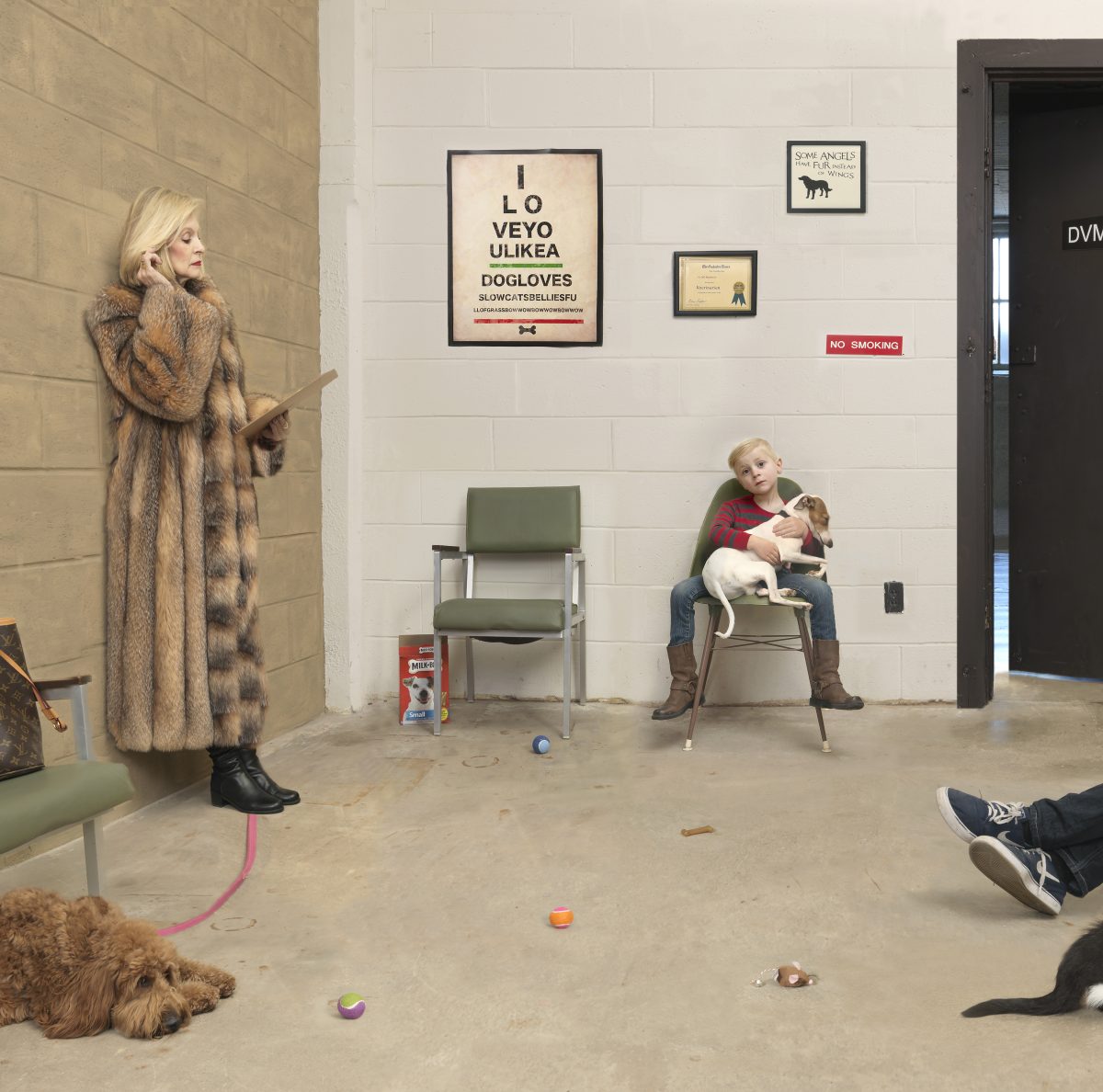 A woman in a fur coat stands beside a man in dark shoes with legs stretched out on the floor, while a young boy sits holding a small dog in a green chair. Dogs and toys scatter across a concrete floor in a clinical waiting room with white brick walls, framed dog-themed posters, and a "No Smoking" sign.