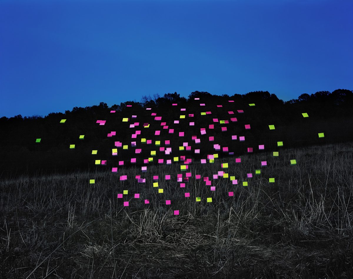 Nighttime landscape featuring a hillside silhouetted against a deep blue sky, with the dark treeline and dry grass rendered in muted tones. Hundreds of neon pink and yellow-green fluorescent squares are densely scattered across the composition, creating a glowing constellation of color that contrasts dramatically with the monochromatic surroundings.