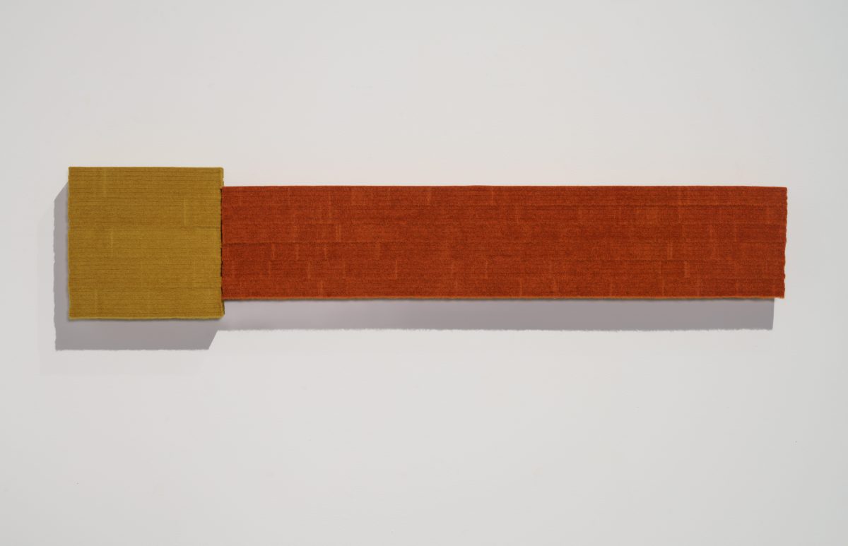Two rectangular panels of industrial felt joined horizontally—a smaller ochre-yellow square on the left and a longer rust-orange rectangle extending to the right—mounted on a white wall, casting soft shadows beneath.