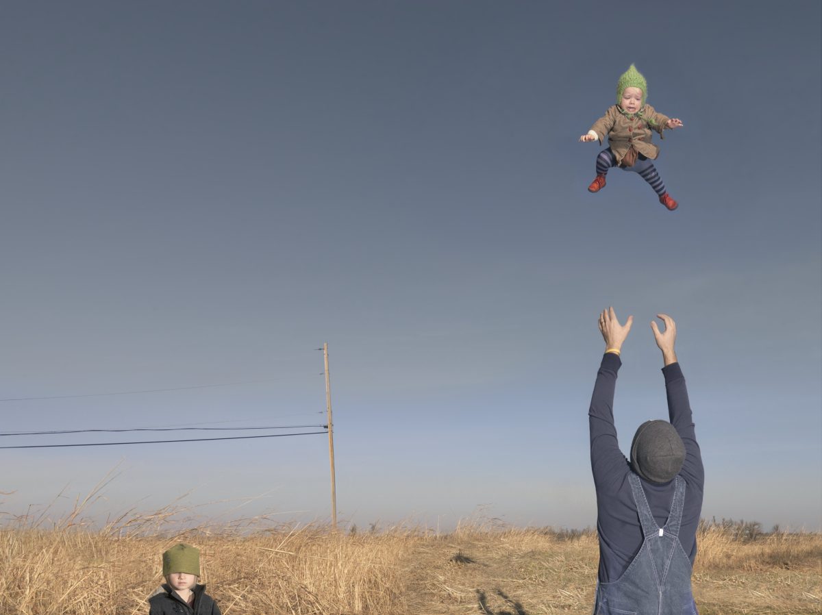 An adult wearing a dark jacket and green hat tosses a small child in a green knit hat high into a clear blue sky above a vast, dry grass field. Another child in a green hat stands in the foreground as the adult's arms reach upward to catch the airborne child, with a utility pole visible on the horizon.