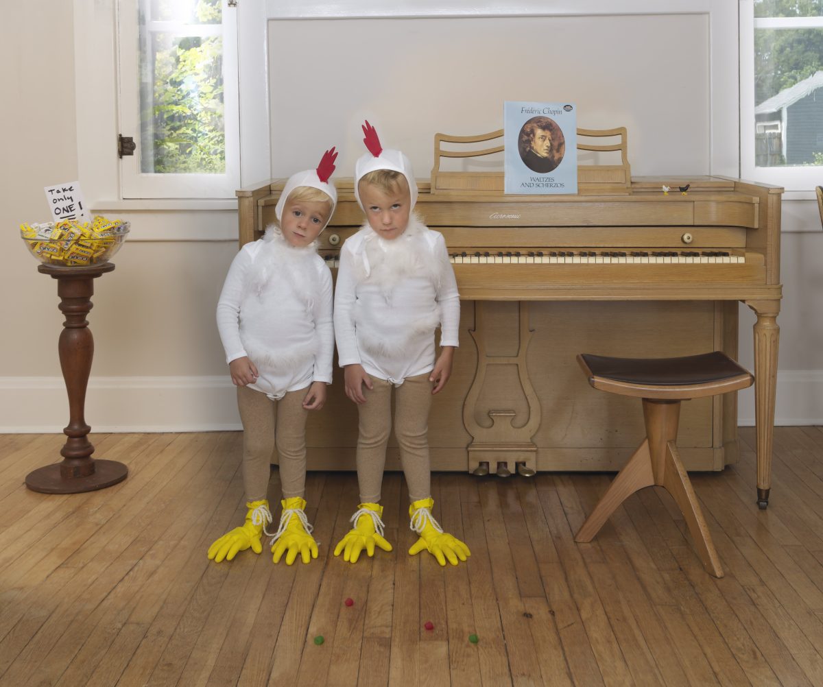 Two young children dressed in white long-sleeved shirts and tan leggings stand before an upright piano, wearing headbands with red feathers and yellow chicken feet on their hands and feet. A wooden pedestal table holding a bowl of candy and a framed Chopin poster sits to the left, while sheet music and a piano bench are visible in the brightly lit room with large windows overlooking greenery.