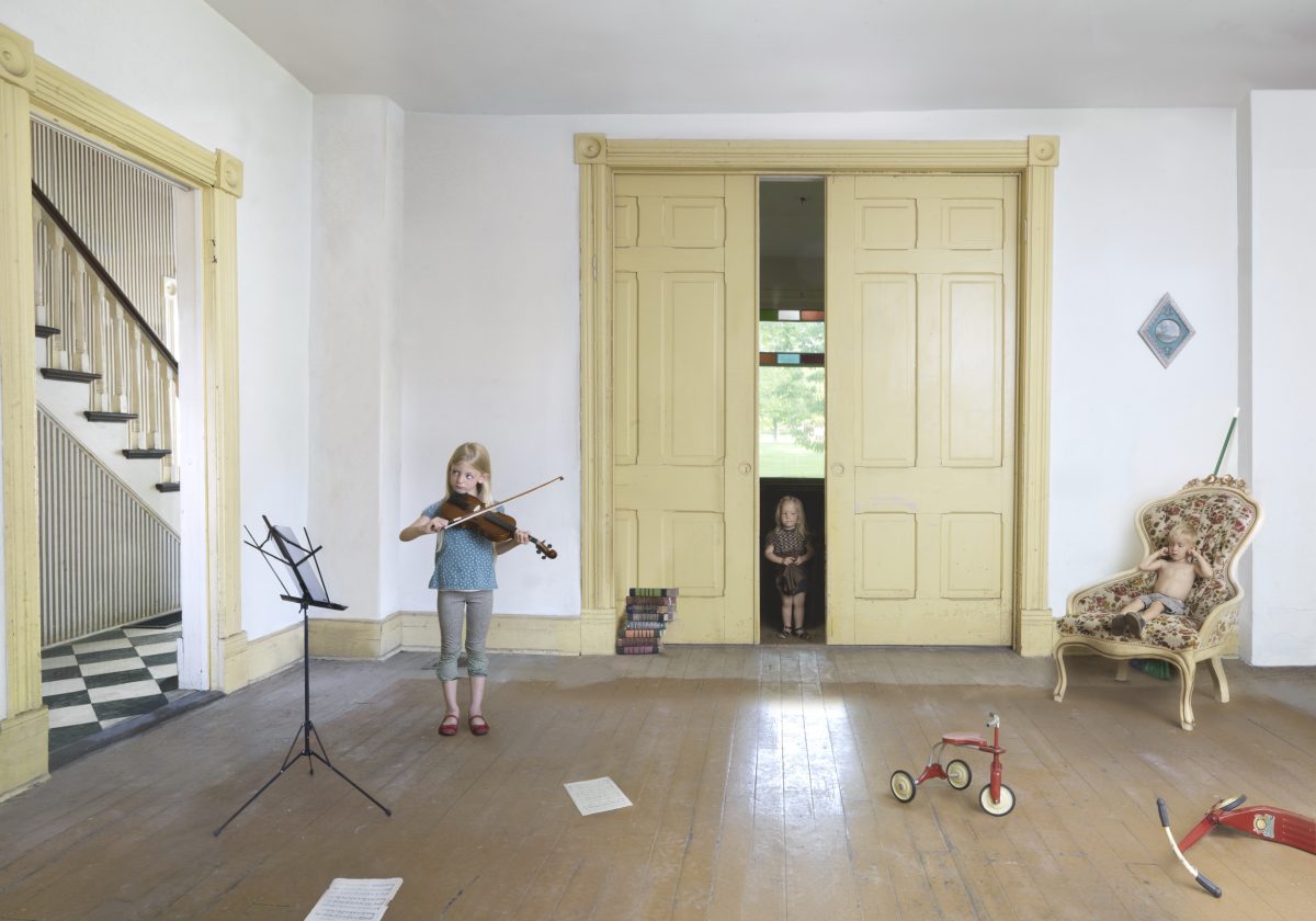 A young girl plays violin in a spacious, cream-colored room with pale yellow trim and large double doors. The bright, formally appointed space contains period furniture, a red tricycle, and scattered musical notation sheets on the wooden floor, while another child peers out from behind the closed doors.